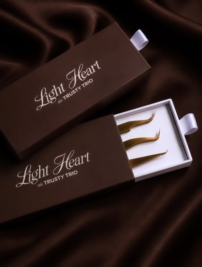 Light Heart Lashes Trusty Trio lash extension tweezer set showing three gold tweezers — the Maddi, Sweetie, and Katy — in an open dark brown keepsake gift box on a satin background